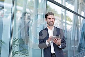 Cheerful and successful businessman works outdoors near the office holds a tablet, smiles reads good news