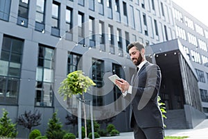 Cheerful and successful businessman works outdoors near the office holds a tablet, smiles reads good news