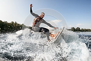 Cheerful surfer riding foaming river wave from motorboat at sunny day.