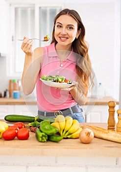 Cheerful svelte young woman eating vegetable salad in home kitchen