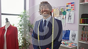 A cheerful tailor with a beard stands confidently in a colorful atelier surrounded by fabrics and a mannequin photo