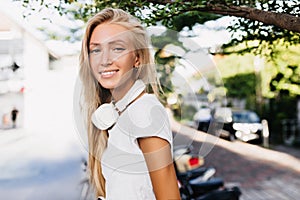 Cheerful tanned lady in headphones spending time outdoor in warm sunny day. Caucasian girl in white