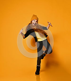 Cheerful teen girl with long red hair in knitted hat, yellow glasses and t-shirt is dancing cool like a hip-hop rap star