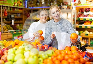 Cheerful teen girl with mother choosing sweet oranges in greengrocery