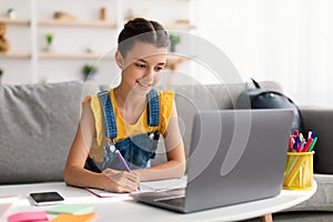 Cheerful teen sitting at desk, using laptop, drawing in copybook