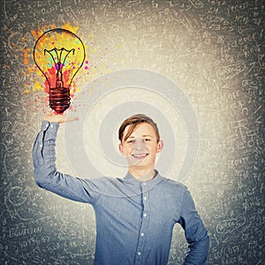 Cheerful teenage boy keeps arm up holding a colorful light bulb as solving hard mathematics calculation, formula and equations.