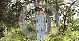 A cheerful teenage girl with curly hair stands in a clearing between green trees in summer forest.