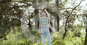 A cheerful teenage girl with curly hair stands in a clearing between green trees in summer forest and has a pleasure