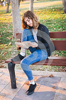 Cheerful teenager girl reading book on the bench at autumn park