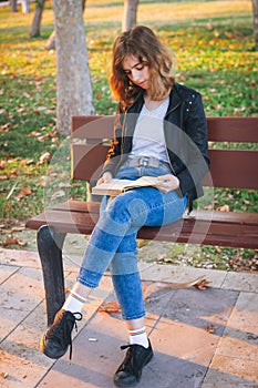 Cheerful teenager girl reading book on the bench at autumn park