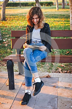 Cheerful teenager girl reading book on the bench at autumn park