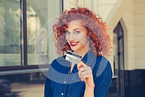 Cheerful toothy smile woman holding showing credit card near bank office store shopping mall outdoors. Curly red head happy girl