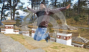 Cheerful traveler man backpacker wearing a biker jacket, jumping hand looking Dochula Dzong architecture of buddhist temple in