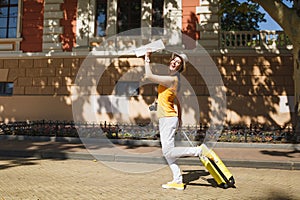 Cheerful traveler tourist woman in yellow summer casual clothes, hat with suitcase city map walk in city outdoor. Girl