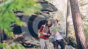 Cheerful travelers are looking at map standing in wood near rocks discussing way and looking around