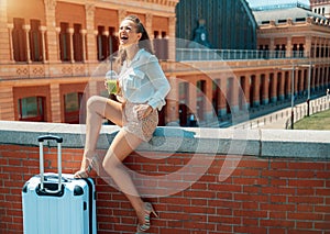 Cheerful traveller woman with green smoothie sitting on parapet