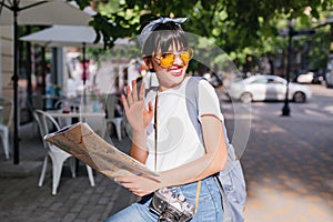 Cheerful trendy girl with camera waving hand in silver ring and smiling, looking away. Outdoor portrait of charming