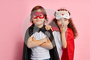 Cheerful two children in cloaks and mask isolated over pink background