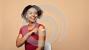 Cheerful vaccinated black woman showing arm with bandage after coronavirus vaccine injection on beige background