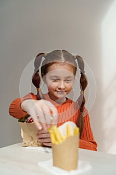 Cheerful white girl eating fries and sandwich in a restaurant. Portrait of pretty elementary age kid eats fast food for lunch in