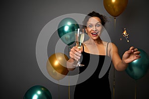 Cheerful woman in black evening dress holds a glass of champagne and sparklers, poses against gray background with beautiful