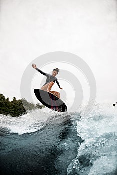 Cheerful woman in black wetsuit effectively jumps on surfboard on wave