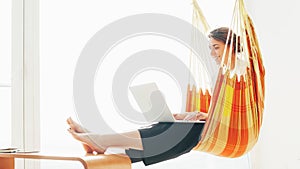 Cheerful woman chilling in hammock with laptop, near big bright window