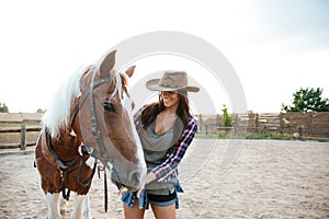 Cheerful woman cowgirl standing and taking care of her horse