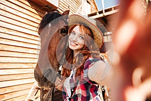 Cheerful woman cowgirl standing taking selfie with horse on farm