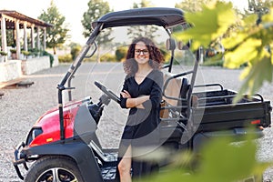 Cheerful woman with curly hair in black dress holding in hands a laptope, poses near vehicle and smiling.