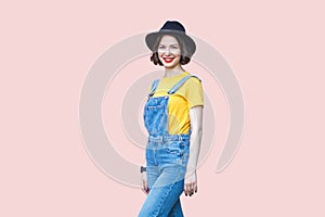 Cheerful woman in denim overalls, yellow T-shirt and black hat, looking at camera with toothy smile.