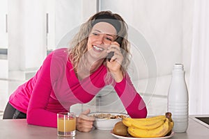 Cheerful woman eating cereal with fruit and milk for breakfast while talking on the phone.