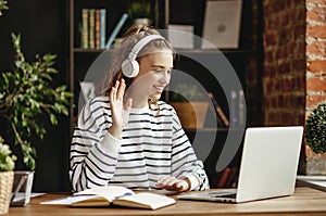 Cheerful woman in headphones greeting friend while talking on laptop at home