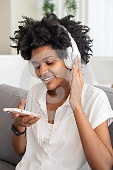 Cheerful woman in headphones sitting on sofa and using phone