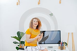 Cheerful woman holding phone in hands sitting on her work desk, smiling