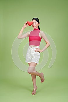 cheerful woman holding red heart while standing with hand on hip isolated on green