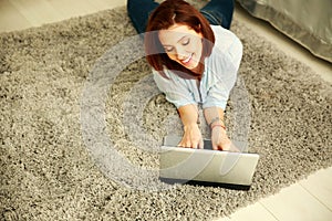 Cheerful woman lying on the carpet and typing on the laptop