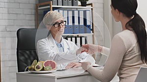 Cheerful woman nutritionist giving nutrition plan to happy female patient at office, grateful lady shaking doctor's hand