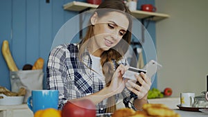 Cheerful woman paying bills online using smartphone and credit card while have breakfast in the kitchen at home