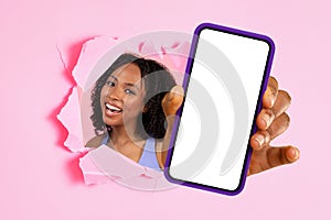 Cheerful woman peeking through a torn pink paper wall presenting a smartphone with a blank white screen