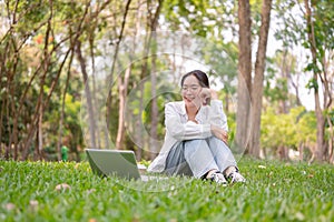 A cheerful woman sits on the grass in a park, enjoying a moment of relaxation with her laptop nearby