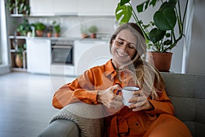 Cheerful woman with smile recalls happy moment from past while sits with cup of tea on sofa at home.