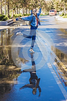 A cheerful woman stands on one leg in a dancing pose on a wet alley in the park. Mirror reflection in a puddle. Dance on