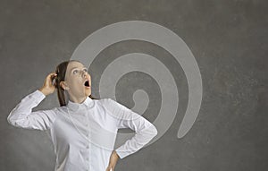 Cheerful woman with a surprised expression looks up while standing on a gray background.