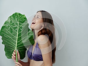 cheerful woman in a swimsuit flatters a big green leaf in her hands  background