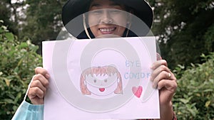 Cheerful woman take off a facial mask with smile stay outdoor and holding a health campaign goodbye coronavirus picture in hands.