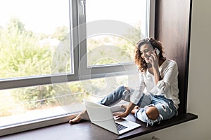 Cheerful woman talking phone and sitting on windowsill with laptop computer at home