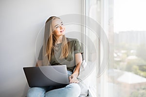 Cheerful woman talking phone and sitting on windowsill with laptop at home