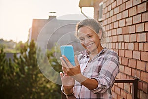 Cheerful woman using smartphone, making selfie standing on the balcony of a country side house on sunny warm summer day