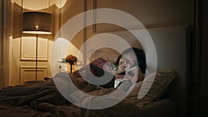 Cheerful woman watching tablet in bed. Happy female laughing movie in evening.